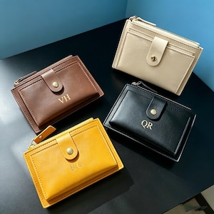 May include: Four rectangular wallets in beige, brown, black, and yellow, each featuring a snap-button flap closure, a top zipper, and gold-toned hardware. The brown wallet is embossed with VH, the black with QR, and the yellow with RG.