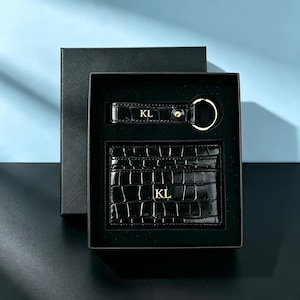 May include: A black crocodile-embossed leather card holder and a matching keychain set presented in a black gift box. Both items feature the gold-embossed initials KL. The keychain includes a gold-toned metal ring and a single gold-toned stud.