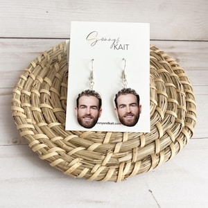 May include: A pair of silver dangle earrings with a photo of a man's face on each earring. The earrings are on a white card with the text "Sonny & Kait" and the website "sonnyandkait.com".