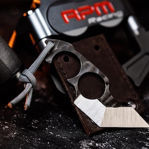 Può includere: Un piccolo coltello in metallo a lama fissa con impugnatura a due fori per le dita e un cordino in paracord grigio. La lama presenta una finitura spazzolata e una punta angolata affilata. È posizionato contro un fodero in pelle marrone e un pezzo RPM.