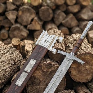 May include: A double-edged sword with a patterned Damascus steel blade, a dark wood handle, and a matching wooden scabbard. The scabbard features a silver-toned metal locket engraved with a Jerusalem cross symbol and a rectangular metal plate.