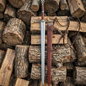 May include: A steel longsword with a twisted brown wooden grip, a silver crossguard, and a matching pommel. Beside the sword is a brown leather scabbard featuring silver metal accents and an adjustable brown leather shoulder strap harness.
