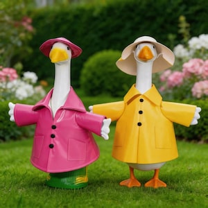 May include: Two goose figurines standing side by side. The left goose wears a bright pink raincoat and matching hat. The right goose wears a yellow raincoat and a beige sun hat. Both geese have white bodies, orange beaks, and orange feet.