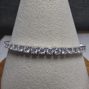 May include: A silver-toned tennis bracelet featuring a continuous row of round, clear, faceted stones set in individual metal prongs. The bracelet is displayed horizontally, showcasing the uniform size and sparkle of each stone along the band.