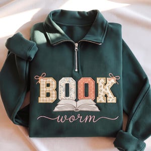 May include: A dark green quarter-zip sweatshirt featuring the word BOOK in large, patterned block letters with small bows on the outer letters, a graphic of an open book below, and the word worm in cursive script underneath the book graphic.
