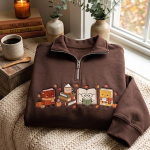 Puede incluir: Una sudadera marrón oscuro con cremallera de un cuarto y un bordado de libros antropomórficos, una pila de libros con una taza y pequeñas hojas de otoño naranjas. El diseño incluye personajes de libros sonrientes, uno con gafas, y calabazas.