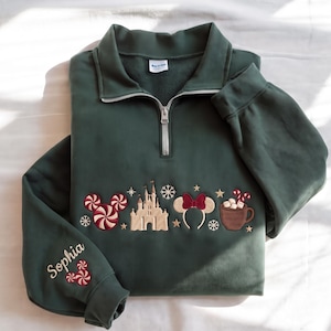 Puede incluir: Una sudadera verde oscuro con cremallera de un cuarto que presenta iconos navideños bordados, incluyendo una cabeza de Mickey de menta, un castillo, una diadema de orejas de Minnie y una taza de cacao. La manga tiene bordado el nombre Sophia.