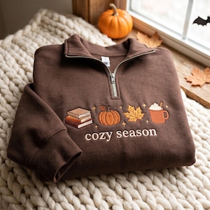 May include: A dark brown quarter-zip sweatshirt featuring embroidered autumn-themed icons including a stack of three books, a pumpkin, a maple leaf, and a mug with whipped cream. The text "cozy season" is embroidered below the icons in white.