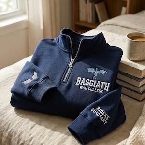 Könnte beinhalten: Ein marineblaues Sweatshirt mit kurzem Reißverschluss, Drachengrafik und dem Schriftzug "BASGIATH WAR COLLEGE" auf der Brust. Der linke Ärmel zeigt ein Flügelsymbol, während auf dem rechten Ärmel der Text "RIDERS QUADRANT" zu sehen ist.
