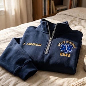 Pode incluir: Uma camisola azul-marinho com fecho de correr de um quarto, com o texto bordado J. ANDERSON no peito direito e um emblema azul da Estrela da Vida com o texto CITY OF RIVERDALE EMS bordado a amarelo no peito esquerdo, com um fecho de correr metálico prateado.
