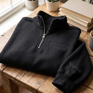May include: A black quarter-zip sweatshirt featuring the embroidered text OCCUPATIONAL THERAPY on the chest and the name Sophia OT embroidered on the sleeve. The garment has a silver-toned metal zipper and ribbed cuffs at the end of the sleeves.