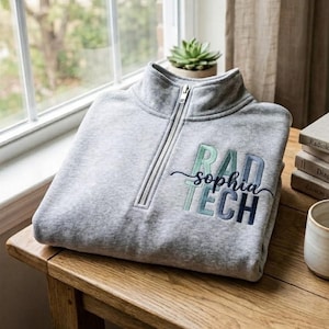 Op de afbeelding: Een grijs gemêleerde trui met een korte rits en geborduurde tekst RAD TECH in verticale blokletters met een kleurverloop van groen naar blauw, en de naam sophia in een zwart cursief lettertype horizontaal over het midden.