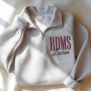 May include: A light gray quarter-zip sweatshirt featuring long sleeves and a collar. The left chest area displays the embroidered text RDMS in a dark pink serif font, with the name Lauren embroidered below it in a black cursive script.