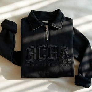 Pode incluir: Uma camisola preta com fecho de um quarto, gola dobrada e fecho de metal prateado. A parte da frente apresenta o texto bordado BCBA em letras grandes de bloco, com as palavras BOARD CERTIFIED BEHAVIOR ANALYST bordadas por baixo.