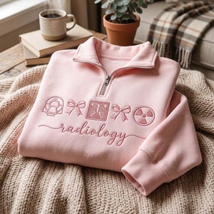 May include: A light pink quarter-zip sweatshirt featuring embroidered designs of a CT scanner, a bow, an X-ray of a rib cage, another bow, and a radiation symbol. The word radiology is embroidered in a cursive script font below the icons.