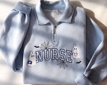 May include: A light blue quarter-zip sweatshirt featuring the word NURSE embroidered in a blue patterned font. The design includes two white ghost figures wearing nurse caps, black spider webs, and small black bats across the chest area.