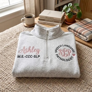 May include: A light grey quarter-zip sweatshirt featuring embroidered text. On the left, it reads Ashley M.S.-CCC-SLP in pink and black. On the right, a circular design reads Speech Language Pathologist with SLP in the centre and a small pink heart.