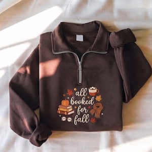 以下が含まれることがあります： 「all booked for fall」という文字が刺繍されたダークブラウンのハーフジップスウェットシャツ。デザインには、本の山、カボチャ、ホットココアのマグカップ、スカーフ、眼鏡、オレンジと茶色の秋の葉が描かれています。