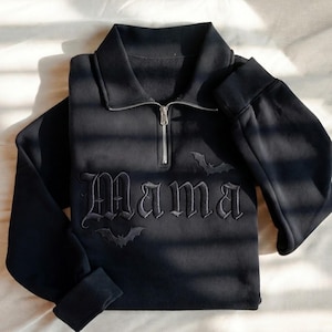 Könnte beinhalten: Ein schwarzes Langarm-Sweatshirt mit Viertelreißverschluss, das auf der Brust das Wort Mama in gotischer Schrift gestickt hat. Das Design enthält zwei kleine gestickte Fledermaussilhouetten über und unter dem Text sowie einen silberfarbenen Reißverschluss.