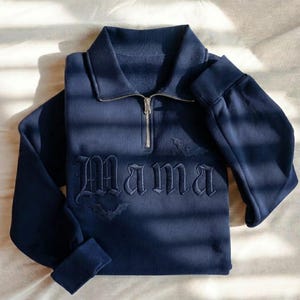 May include: A navy blue quarter-zip sweatshirt featuring the word Mama embroidered in a gothic font across the chest. The design includes two small embroidered bat silhouettes, one above and one below the text, with a silver metal zipper.