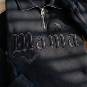 May include: A black quarter-zip sweatshirt featuring the word Mama embroidered in a gothic font across the chest. The design includes two small embroidered bats, one positioned above the text and one below, with a silver-toned metal zipper.