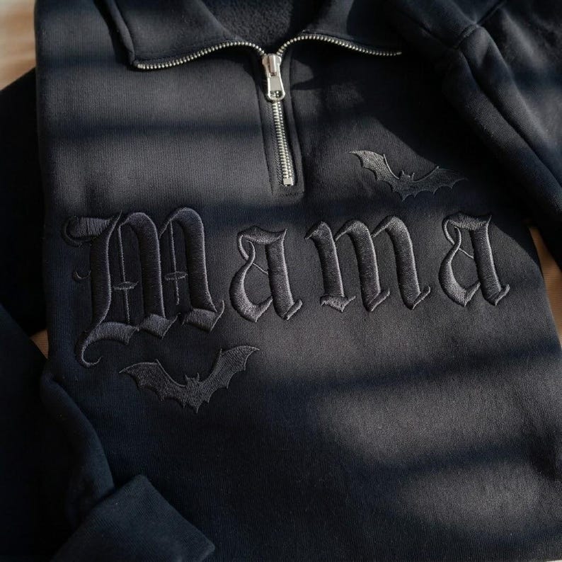 May include: A black quarter-zip sweatshirt featuring the word Mama embroidered in a gothic font across the chest. The design includes two small embroidered bats, one positioned above the text and one below, with a silver-toned metal zipper.