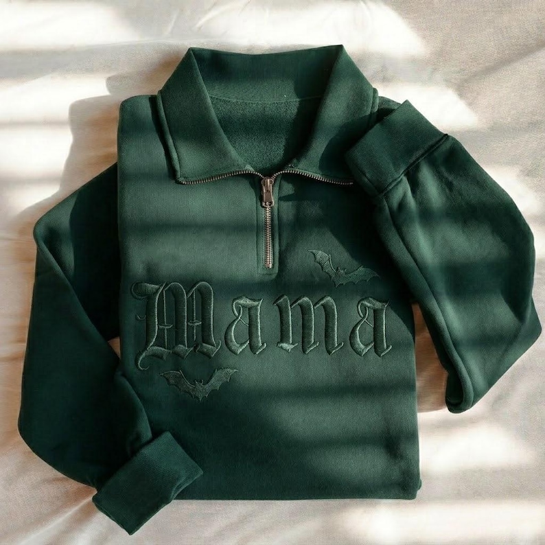 May include: A dark forest green quarter-zip sweatshirt featuring long sleeves, a high collar, and a metal zipper. The front is embroidered with the word "Mama" in a gothic font, accompanied by two small bat silhouettes positioned above and below.
