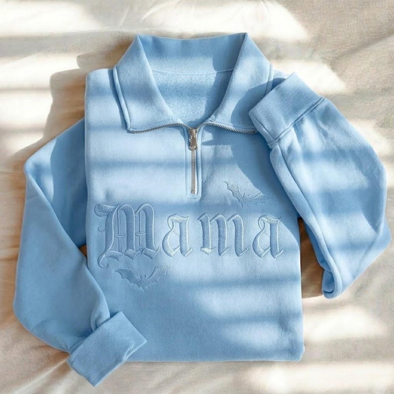 May include: A light blue quarter-zip sweatshirt featuring a fold-over collar and long sleeves. The front displays the word "Mama" embroidered in a gothic font, accompanied by two small embroidered bat silhouettes positioned above and below the text.