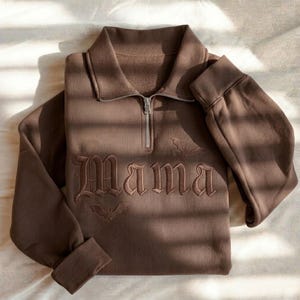 May include: A dark brown quarter-zip sweatshirt featuring the word Mama embroidered in a gothic font across the chest. The design includes small embroidered bat silhouettes above and below the text, with a silver-toned metal zipper pull.