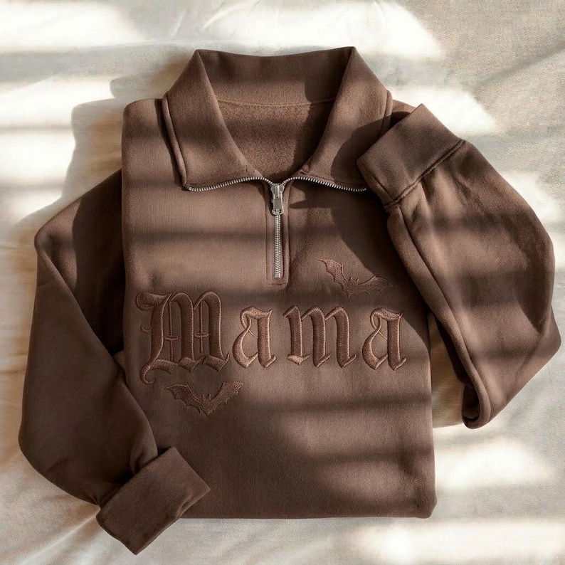 May include: A dark brown quarter-zip sweatshirt featuring the word Mama embroidered in a gothic font across the chest. The design includes small embroidered bat silhouettes above and below the text, with a silver-toned metal zipper pull.