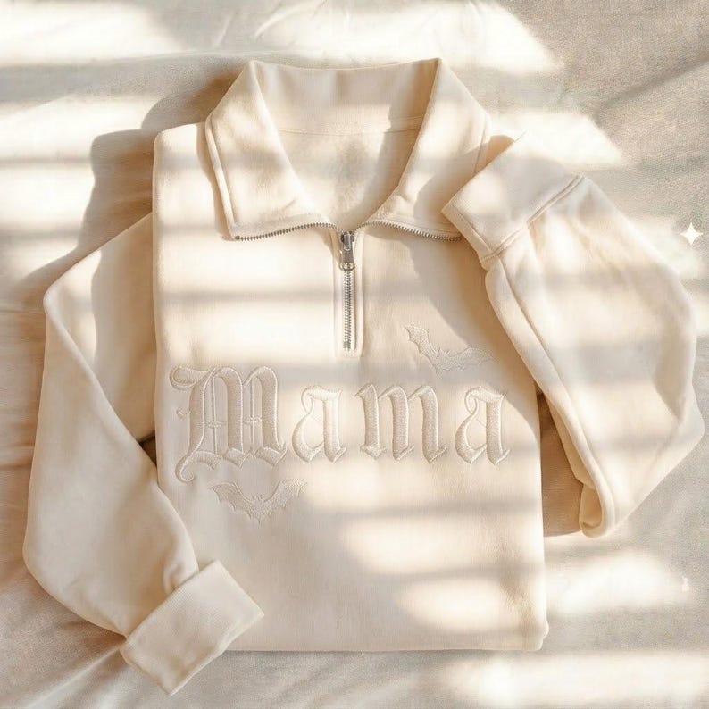 May include: A cream-colored quarter-zip sweatshirt featuring the word Mama embroidered in a gothic font across the chest. The design includes small embroidered bat motifs positioned above and below the text, with a silver metal zipper pull.