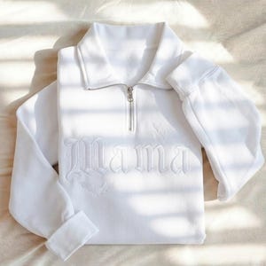 May include: A white long-sleeve quarter-zip sweatshirt featuring the word Mama embroidered in a gothic font across the chest. The design includes small embroidered bat silhouettes integrated with the lettering on a soft, solid white fabric.
