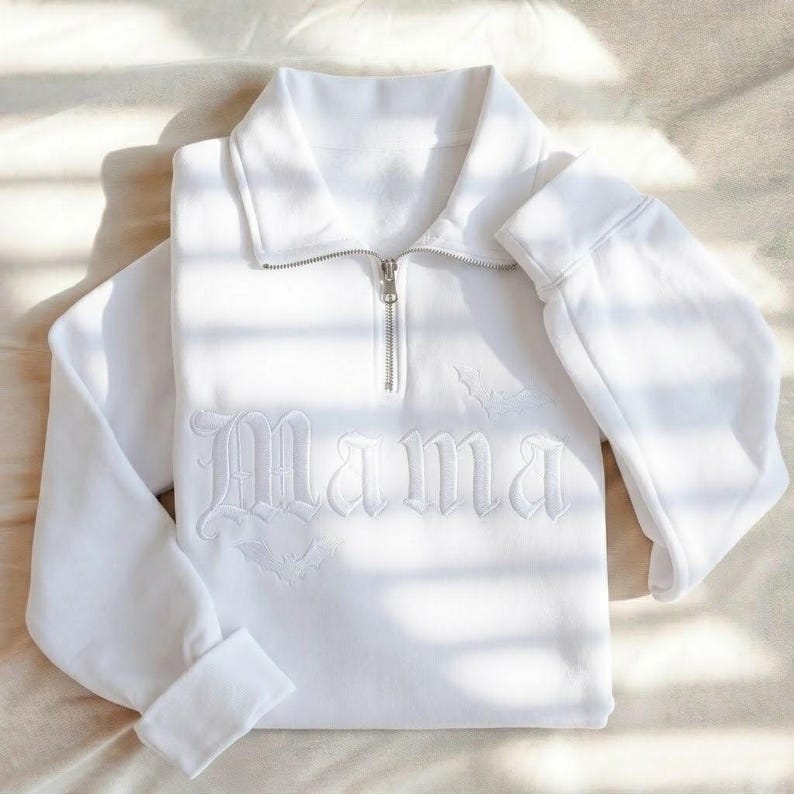 May include: A white long-sleeve quarter-zip sweatshirt featuring the word Mama embroidered in a gothic font across the chest. The design includes small embroidered bat silhouettes integrated with the lettering on a soft, solid white fabric.
