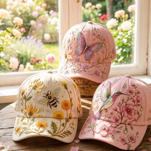 Puede incluir: Tres gorras de béisbol con bordados detallados. La gorra superior es rosa claro con una mariposa y motivos florales. La gorra inferior izquierda es crema con una abeja y dientes de león. La gorra inferior derecha es rosa con un colibrí y flores.