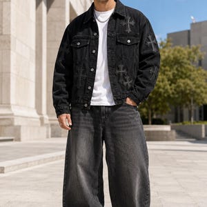 May include: A black denim jacket featuring tonal cross-shaped patches, a white crew-neck t-shirt, and loose-fitting charcoal gray denim jeans. The jacket has a button-front closure, two chest pockets, and long sleeves with additional cross-shaped patches.