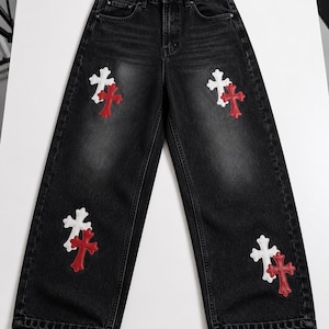 May include: Black denim jeans featuring a high-waisted design with a button and zipper fly closure. The front of the legs is decorated with four distinct cross-shaped patches, each consisting of a white cross layered over a red cross.