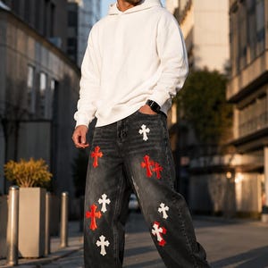 Peut inclure: Un sweat-shirt à capuche blanc associé à un jean en denim gris foncé à la coupe ample. Le jean présente un motif répétitif de croix brodées en contraste, de couleurs rouge et blanche, réparties sur le devant des jambes du pantalon.
