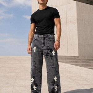 May include: A black short-sleeved t-shirt and loose-fitting charcoal gray denim jeans featuring white and black cross-shaped patches and decorative stitching patterns on the legs, paired with black and white athletic sneakers.
