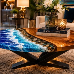 Peut inclure: Une table basse ronde présentant une surface en bois naturel d'un côté et un design océanique en résine bleue avec des vagues blanches et des galets incrustés de l'autre. La table repose sur une base en métal noir en forme de X.