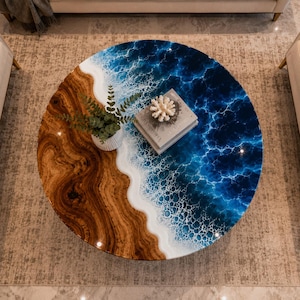 Op de afbeelding: Een ronde salontafel met een sectie van natuurlijk houtnerf aan de ene kant en een sectie van glanzende blauwe hars aan de andere kant, die een oceaankustlijn nabootst met witte schuimdetails. Een kleine plant in pot en een boek liggen bovenop.