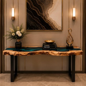 Pode incluir: Uma mesa consola com um tampo de madeira natural de borda viva com uma incrustação central de resina azul, apoiada por duas pernas de metal preto em forma de U. A mesa contém um vaso escuro com flores brancas, uma tigela e dois livros pretos.