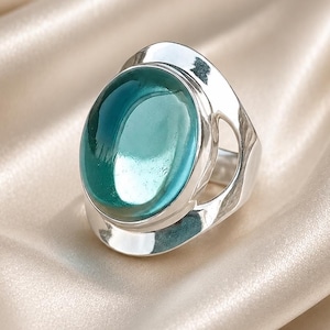Op de afbeelding: Een zilveren ring met een grote, ovale, doorschijnende blauwgroene steen gezet in een gepolijste metalen band. De ring heeft een glad, reflecterend oppervlak en een breed, opengewerkt ontwerp aan de zijkanten dat de centrale edelsteen omlijst.