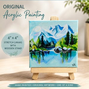 May include: An original acrylic painting on a 4 inch by 4 inch stretched canvas, displayed on a small wooden stand. The artwork depicts a landscape with blue mountains, green trees, and a reflective body of water. The initials VM are in the corner.