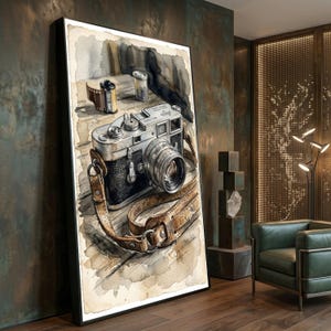 May include: A framed watercolor painting depicting a vintage Leica M3 camera with a brown leather strap resting on a wooden surface. Beside the camera sits a small film canister. The artwork features muted tones of brown, gray, and black ink.