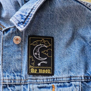 May include: A rectangular black embroidered patch featuring a white crescent moon with a face, surrounded by yellow clouds and a small star, with the text "the moon" in yellow thread, attached to a light blue denim jacket with metal buttons.