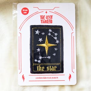 May include: A rectangular iron-on embroidered patch featuring a black background with a large gold star at the centre, surrounded by smaller silver stars and constellations. The text "the star" is embroidered in gold at the bottom of the patch.