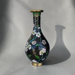 May include: A black cloisonné vase with a narrow neck and a bulbous body, featuring a gold-toned rim and base. The surface is decorated with a repeating gold wire pattern and hand-painted white and pink cherry blossom flowers with green leaves.