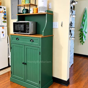 May include: A dark green wooden kitchen cabinet with a natural wood countertop, a single drawer with two black pulls, and two lower doors with vertical paneling. The cabinet features a hutch top with an open shelf and the text HandyByMariam.
