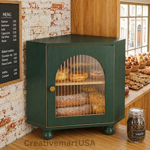 May include: A dark green corner cabinet with a fluted glass arched door and a small gold knob. The cabinet features two interior shelves holding various baked goods, including pastries and loaves of bread, and stands on four rounded feet.