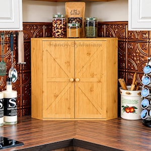 May include: A light brown bamboo corner cabinet with two doors featuring a diagonal cross-brace design and small round wooden knobs. The cabinet has the text Handy ByMariam printed on the front. It is positioned on a dark wood countertop.
